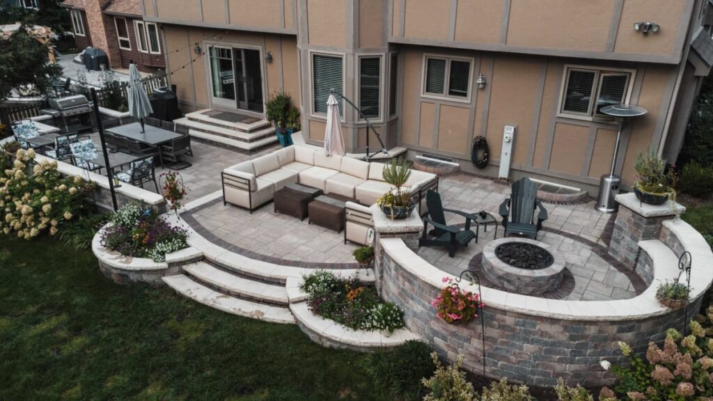 Patio with sectional seating, circular brick fire pit, retaining wall with flowers, and adjacent dining area.